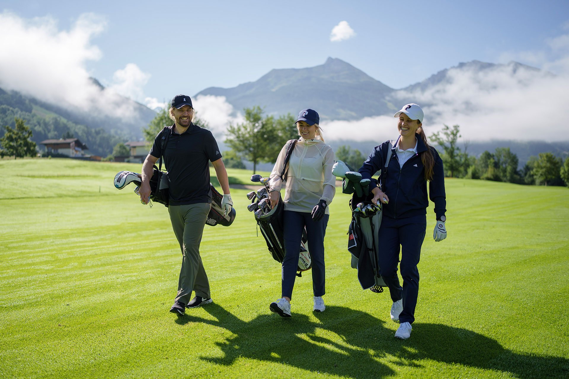 Drei Golfer gehen fröhlich über das grüne Golfplatzgelände, umgeben von Bergen und Wolken im Hintergrund.