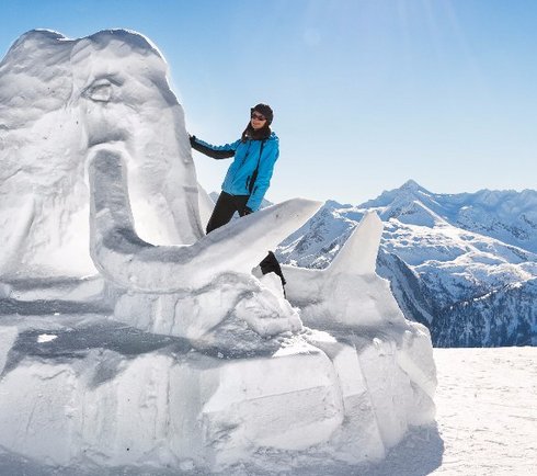 Schneeskulptur Mammut mit Schifahrerin im Vordergrund, Hintergrund blauer Himmel und Berge