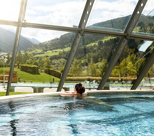 Frau entspannt im Innenbecken der Alpentherme Gastein und blickt durch ein großes Panoramafenster auf die herbstliche Landschaft.