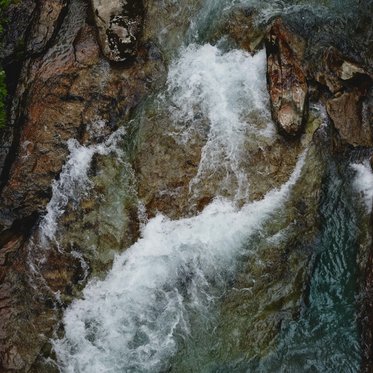 Ein klarer Gebirgsfluss fließt über glatte Steine, während schäumendes Wasser die Felsen umspült und spritzt.
