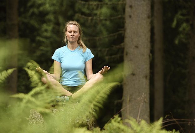 Eine Frau meditiert im Wald, umgeben von hohen Bäumen und grünen Farnen, während sie in Ruhe und Gelassenheit versinkt.