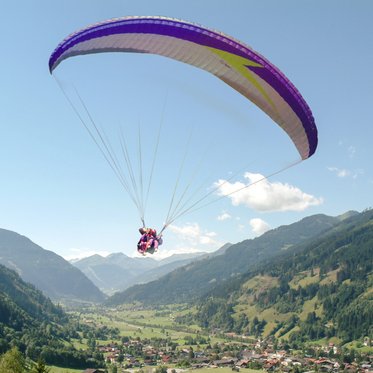 Paragleiter schwebt über Gastein, vor ihm ein Panorama aus grünen Almen, Wäldern und Bergen.