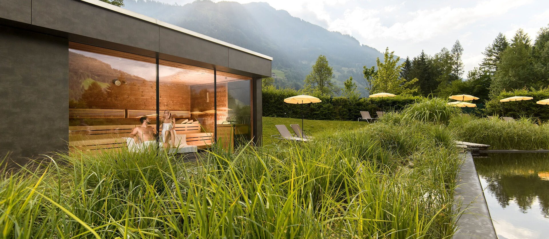 Die Sauna ist aus Holz und Glas, umgeben von hohen Gräsern und Liegen, die zum Entspannen im Freien einladen.