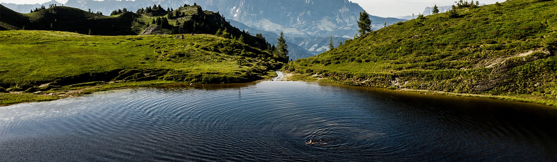 Ein ruhiger Bergsee spiegelt die umliegenden grünen Hügel und majestätischen Berge wider, während eine Person schwimmt.