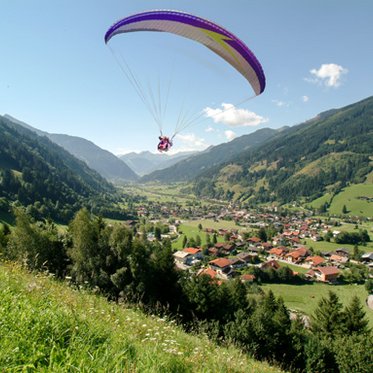 Paragleiter zieht seine Kreise über Gastein. Er ist gerade gestartet, im Tal der Ort Hofgastein, blauer Himmel mit vereinzelten weißen Wolken. 