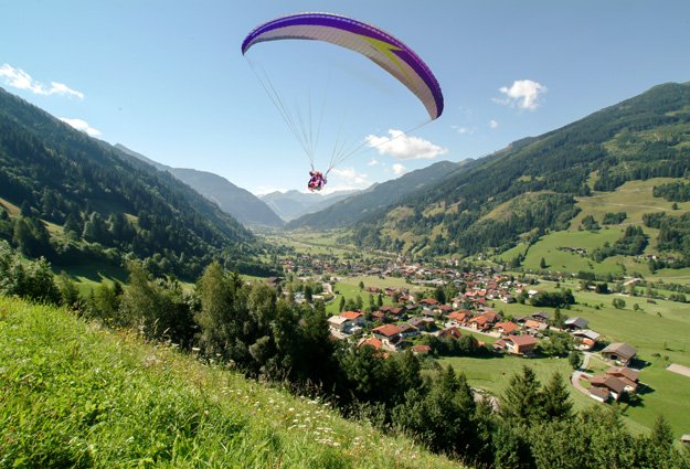 Am linken Bildrand, eine grüne Almwiese, ein kleines Waldstück, ein Paragleiter ist gerade gestartet und schwebt über Gastein hinweg. In der Ferne ein Bergpanorama, an einem schönen Sommerlichen Tag. 