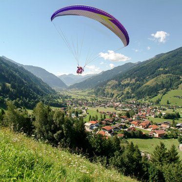 Am linken Bildrand, eine grüne Almwiese, ein kleines Waldstück, ein Paragleiter ist gerade gestartet und schwebt über Gastein hinweg. In der Ferne ein Bergpanorama, an einem schönen Sommerlichen Tag. 