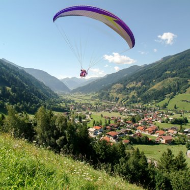 Saftige grüne Wiese am linken Bildrand, ein Paragleiter hat gerade abgehoben. Er schwebt über Gastein, unten sieht man eine Siedlung, ein Bergpanorama in der Ferne.