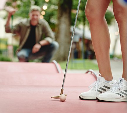 Eine Person steht auf einem Mini-Golfplatz, bereit, den Ball mit einem Golfschläger zu schlagen, während ein Freund im Hintergrund zuschaut.