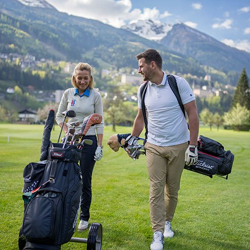 Ein Paar geht entspannt über den Golfplatz, während sie ihre Golftaschen tragen und die schöne Berglandschaft genießen.