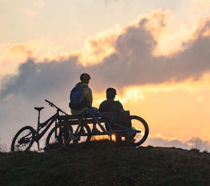 Zwei Personen sitzen bei Sonnenuntergang auf einer Bank auf einem Hügel im Gasteinertal, ihre Mountainbikes stehen daneben.