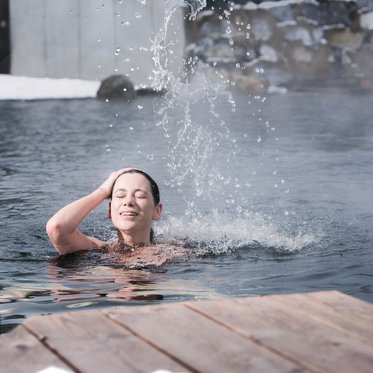 Frau schwimmt im Badeteich im Außenbereich der Alpentherme Gastein, während Dampf vom warmen Wasser aufsteigt.