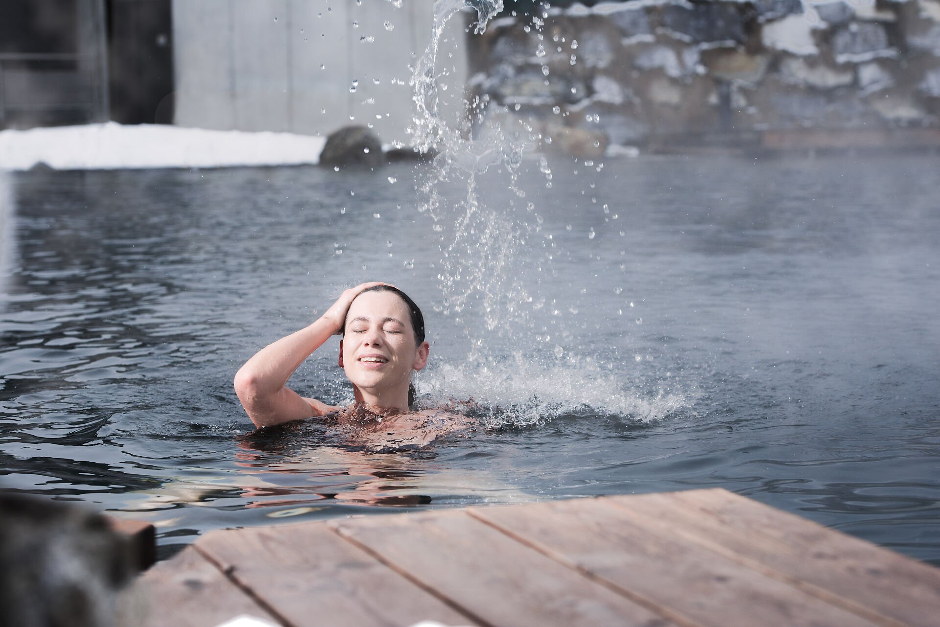 Frau schwimmt im Badeteich im Außenbereich der Alpentherme Gastein, während Dampf vom warmen Wasser aufsteigt.