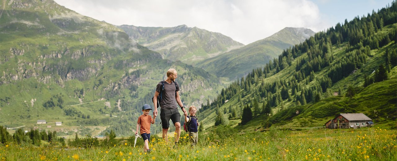 Vater wandert mit zwei Kindern über eine blühende Bergwiese in einer grünen alpinen Landschaft mit Blick auf Berge, Wälder und eine Berghütte.