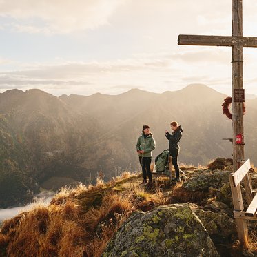 Zwei Wanderinnen stehen bei einem Gipfelkreuz neben einer Bank und genießen den weiten Blick über die Berglandschaft im Gasteinertal.