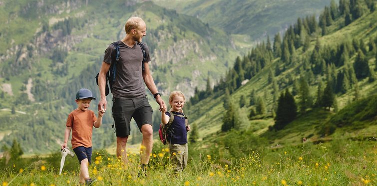 Familie beim Wandern auf einer bunten Bergwiese in den Alpen, umgeben von grünen Berghängen, Wäldern und einer traditionellen Berghütte im Hintergrund.
