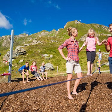 Ein junges Mädchen probiert, unterstütz von seinen Eltern eine Slackline aus. Spielplatz auf der Schlossalm in Gastein.