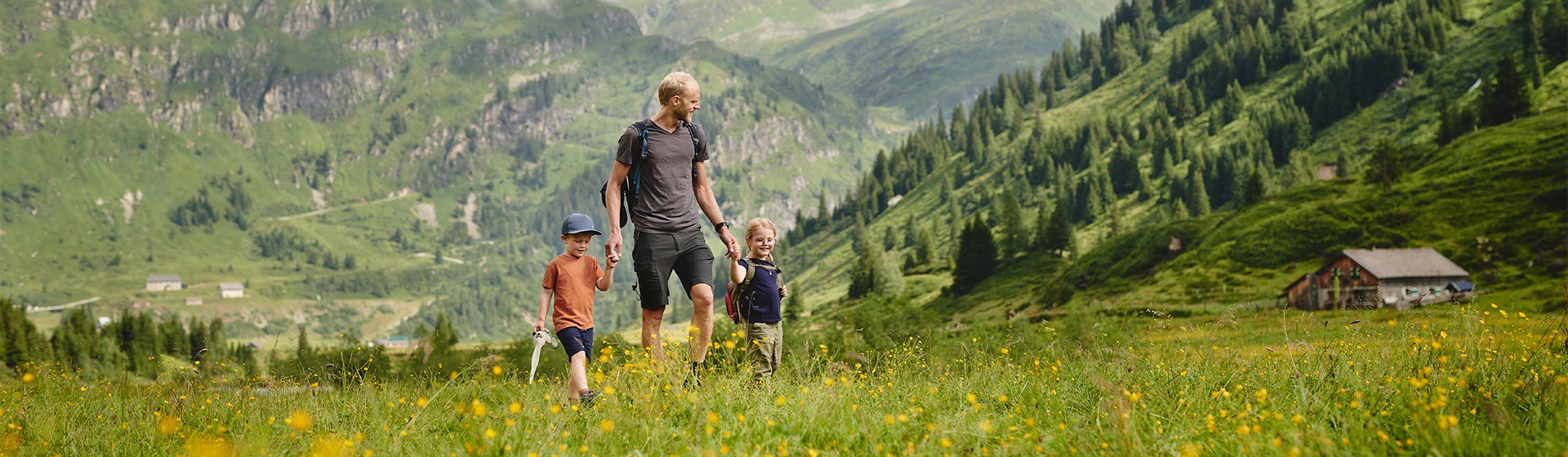 Vater wandert mit zwei Kindern über eine blühende Bergwiese in einer grünen alpinen Landschaft mit Blick auf Berge, Wälder und eine Berghütte.