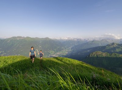 Zwei Personen wandern über einen grünen Bergkamm mit blühender Wiese; im Hintergrund erstreckt sich eine weite Berglandschaft unter klarem Himmel.