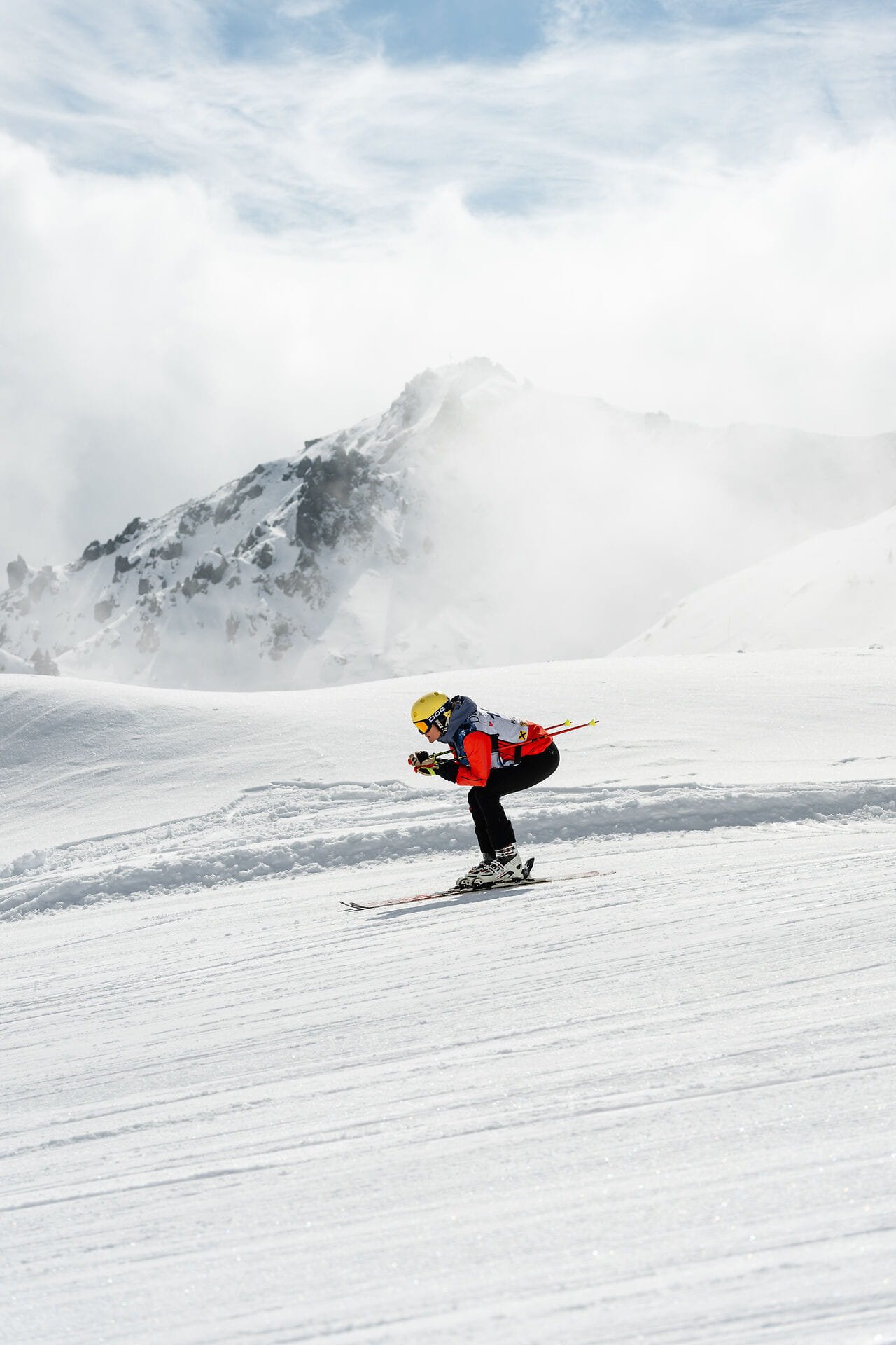 Die Nord 2026 Rennfahrer*in in Hocke auf der Piste, im Hintergrund eine eiskalte Nebelwolke vor einem verschneiten Berg, auf den die Sonne scheint.