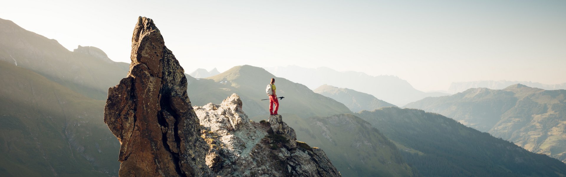Ein Bergsteiger steht auf einem Felsen und blickt über die majestätischen Berge in der Ferne, umgeben von sanften Hügeln.