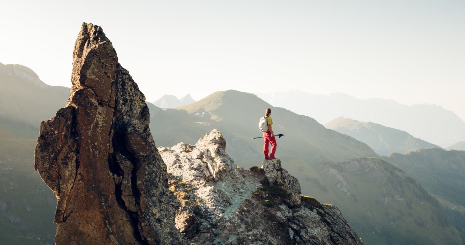 Ein Bergsteiger steht auf einem Felsen und blickt über die majestätischen Berge in der Ferne, umgeben von sanften Hügeln.