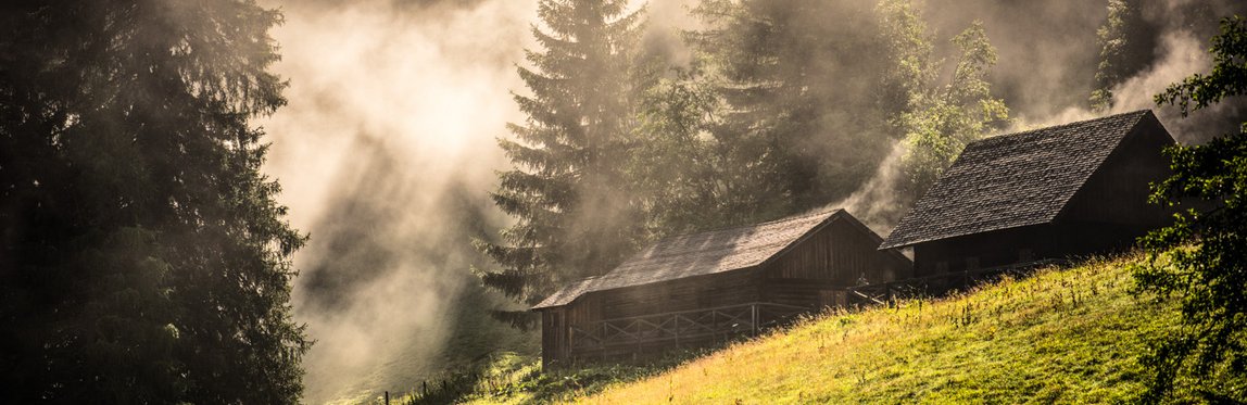 Leichter Nebel steigt auf, an einer Bergwiese, stehen zwei alte Holzhütten, dahinter erste Bäume des Waldes.