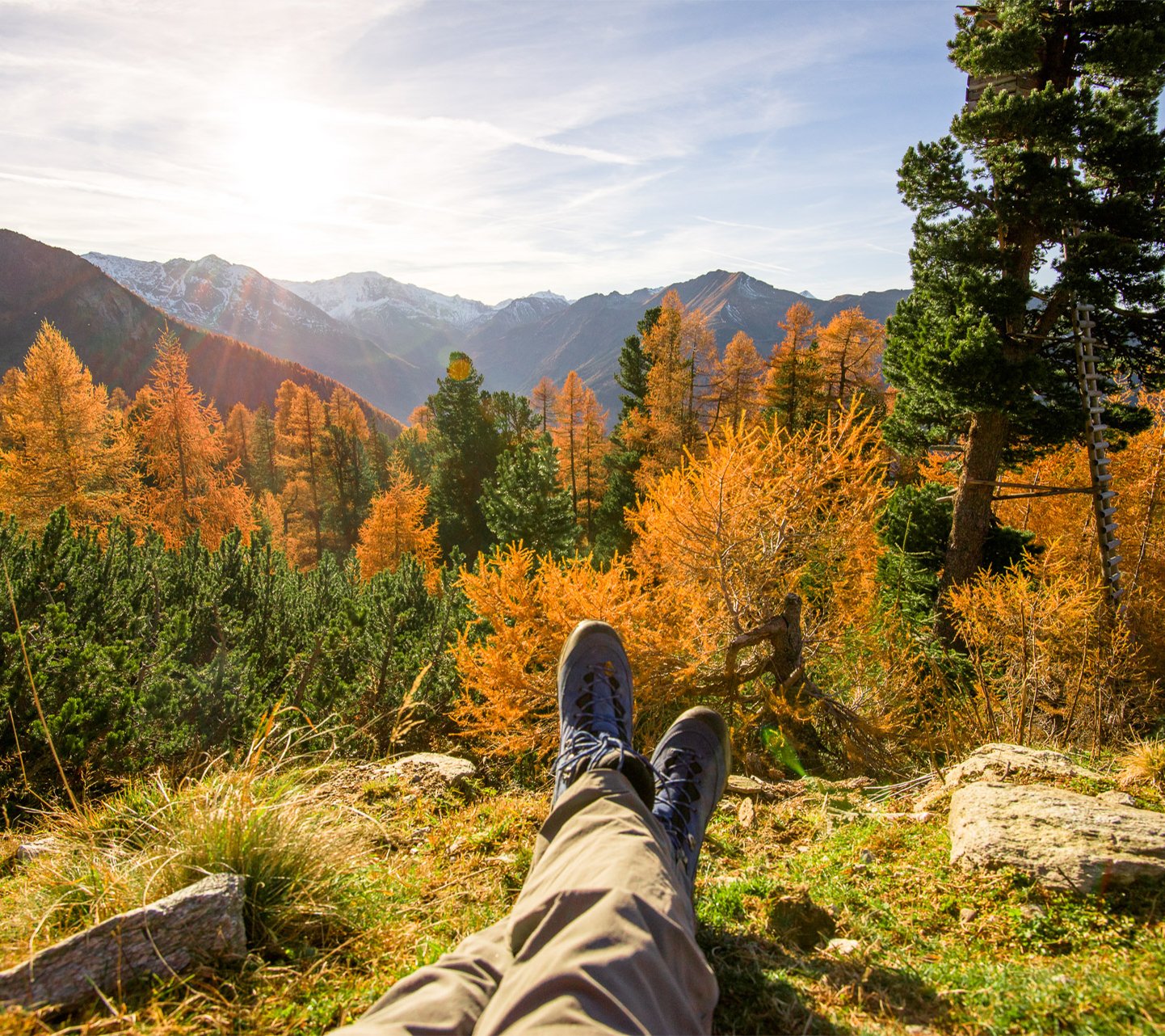 Blick aus der Perspektive einer wandernden Person, die mit ausgestreckten Beinen auf einer Anhöhe sitzt und auf goldene Lärchenwälder sowie die Berggipfel im herbstlichen Gasteinertal schaut.