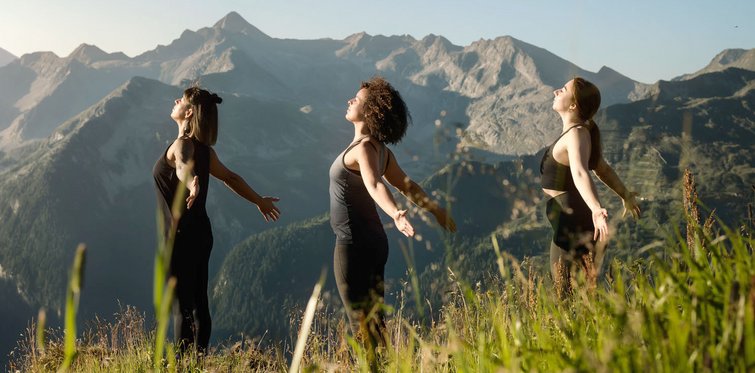 Drei Frauen praktizieren Yoga auf einer sonnigen Bergwiese mit Blick auf eine beeindruckende alpine Berglandschaft und genießen Ruhe, Natur und frische Bergluft.