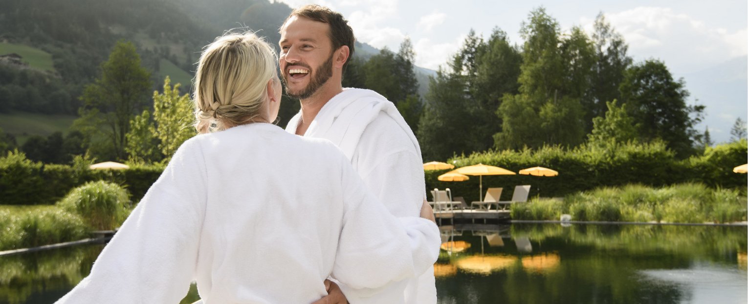 Fröhliches Paar in Bademänteln vor dem Naturbecken einer Therme in Gastein