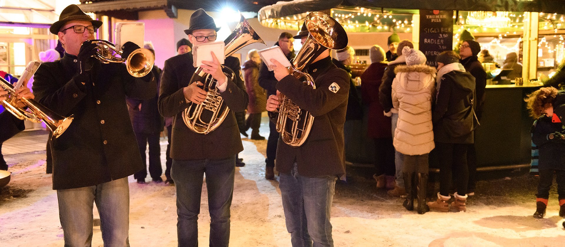 Artvent in Bad Gastein: Musiker spielen Blasinstrumente vor einem festlich beleuchteten Stand, während Besucher in winterlicher Kleidung die vorweihnachtliche Atmosphäre genießen.