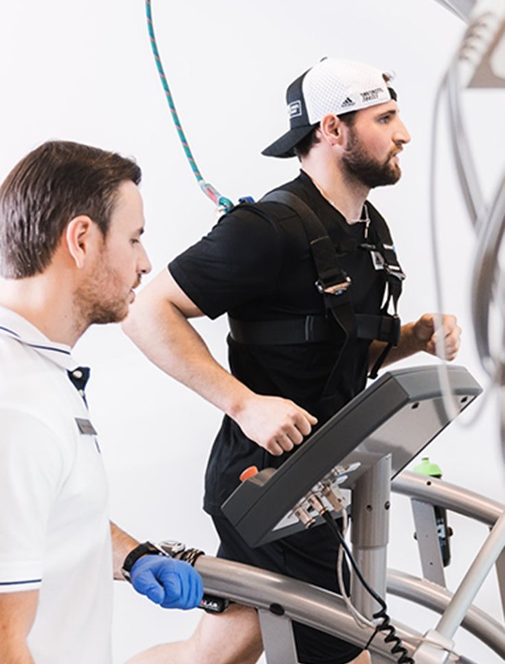 Ein Mann läuft auf einem Laufband, während ein Trainer ihn mit einem speziellen Gurt unterstützt und überwacht.