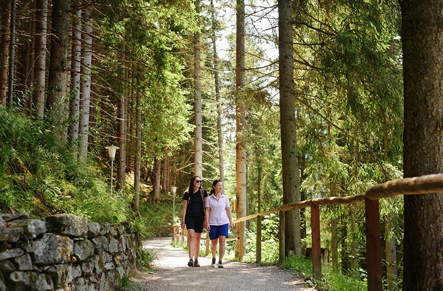 Zwei Frauen spazieren entspannt auf einem schmalen Waldweg, umgeben von hohen Bäumen und üppigem Grün.