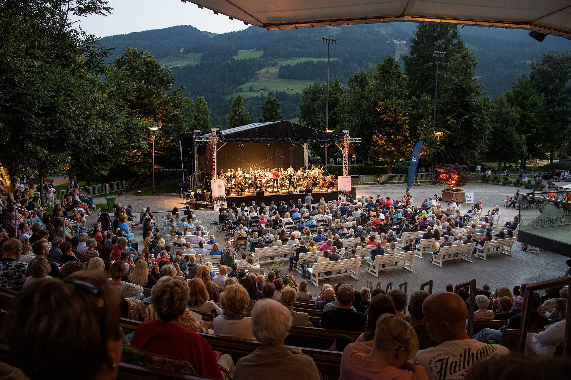 Ein Konzert im Freien zieht viele Besucher an, die auf Holzbänken sitzen und die Musik in der Abenddämmerung genießen.