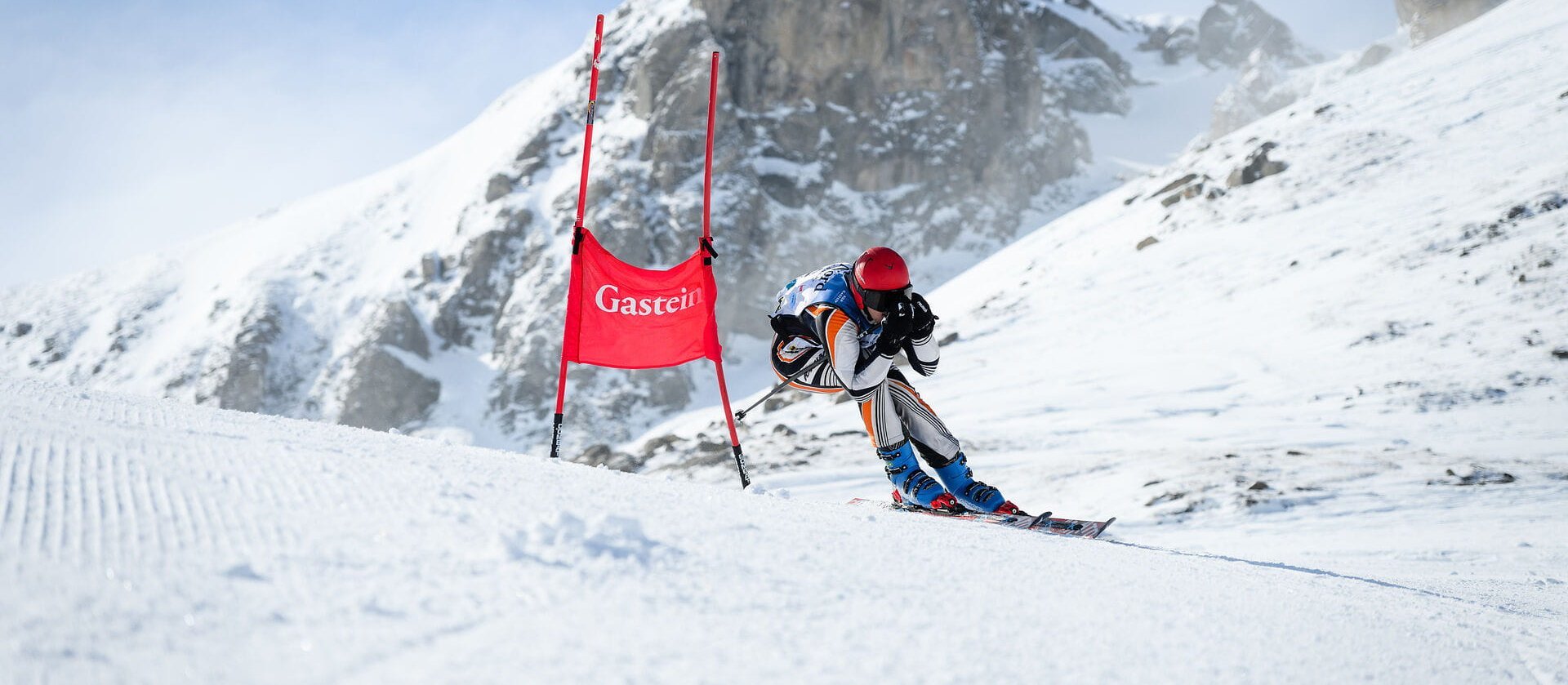 Die Nord 2026 Dynamische Aufnahme von Rennfahrer*in, der/die in tiefer Hocke schenll am Tor vorbeifährt. Dahinter ragt ein verschneiter steiler Berggipfel in die Höhe.