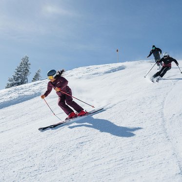 Zwei Skifahrer fahren bei sonnigem Wetter eine präparierte Piste hinunter und ziehen Schwünge im Schnee; im Hintergrund sind verschneite Hänge und vereinzelte Bäume zu sehen.