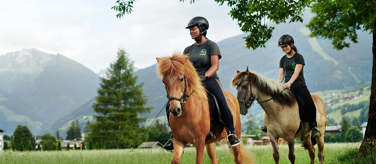 Zwei Reiter*innen auf Pferden reiten durch eine Wiese vor Bergkulisse im Gasteinertal.